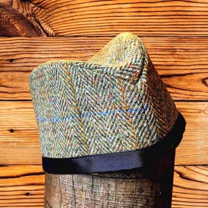 May include: A structured hat featuring a multi-colored herringbone tweed pattern in shades of green, brown, and grey with thin blue and orange overcheck lines. The base of the hat is finished with a solid black grosgrain ribbon trim.