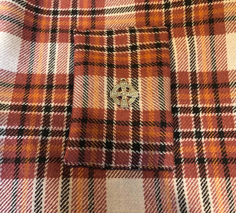 Op de afbeelding: Close-up van een geruite stof in tinten oranje, rood, zwart en wit. Een kleine, zilverkleurige Keltische kruis hanger is op de stof gespeld. Het ruitpatroon is een klassiek ontwerp.