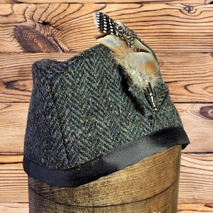 May include: A dark green tweed hat with a black ribbon trim and a decorative feather accent. The hat has a herringbone pattern and is displayed on a wooden form. The feather is a mix of brown, tan, and white.