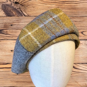May include: A wool beret in a plaid pattern with shades of yellow, gray, and blue. The hat is displayed on a mannequin head. The beret has a classic, rounded shape and a small brim. The plaid design features white and brown accents.