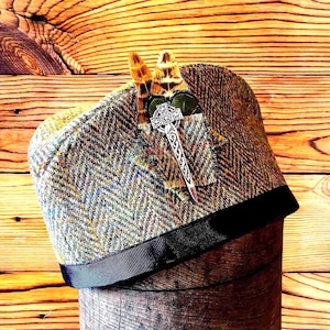 May include: A tweed flat cap with a black ribbon trim, adorned with a silver brooch and two feathers. The cap displays a herringbone pattern in brown, green, and blue. The brooch is shaped like a sword.