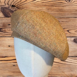 May include: A tan tweed flat cap with a rounded crown and a small, stiff brim. The hat is made of a textured fabric with flecks of red and green. The cap is displayed on a white mannequin head against a wooden background.