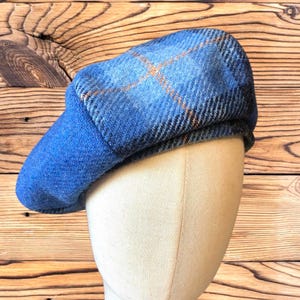 May include: A blue tweed newsboy cap with a plaid pattern in shades of blue and orange. The cap is displayed on a mannequin head, showcasing its shape and design. The hat has a rounded crown and a short, stiff brim.