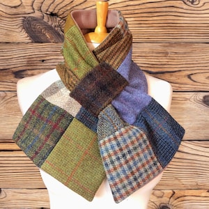 May include: A patchwork scarf constructed from various wool tweed fabrics featuring a mix of herringbone, plaid, and solid patterns in earthy tones of olive green, brown, blue, and purple, with a soft, solid-coloured lining visible at the neck.