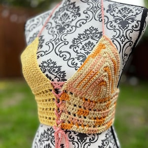 May include: A handmade crochet halter top in shades of yellow, peach, and orange. The top features a lace-up front and a halter neck tie. The crochet work has a textured appearance, and the overall design is bohemian.