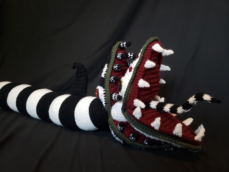 Puede incluir: Un juguete de peluche tejido a crochet con forma de gusano largo y rayado con una boca grande abierta y dientes afilados. El gusano es de rayas blancas y negras y la boca es roja, blanca y verde.