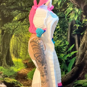 May include: A crochet unicorn hood in white, pink, and blue. The hood has a pink horn and ears, with a blue and white mane. The hood is designed as a costume or accessory.