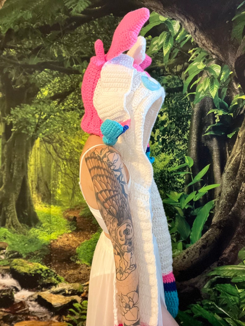 May include: A crochet unicorn hood in white, pink, and blue. The hood has a pink horn and ears, with a blue and white mane. The hood is designed as a costume or accessory.