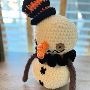 May include: A crocheted snowman with a black and orange hat, a black bow tie, and an orange carrot nose. The snowman has two brown arms and is standing on a wooden surface.