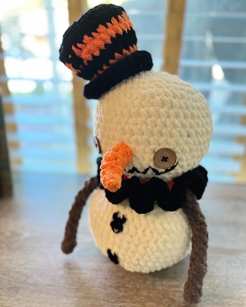 May include: A crocheted snowman with a black and orange hat, a black bow tie, and an orange carrot nose. The snowman has two brown arms and is standing on a wooden surface.