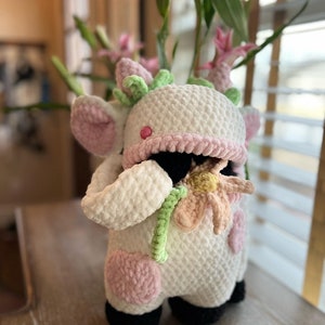 May include: A white crocheted cow plush toy with pink accents and black hooves. The cow is holding a pink and yellow crocheted flower.