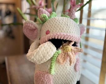 May include: A white crocheted cow plush toy with pink accents and black hooves. The cow is holding a pink and yellow crocheted flower.