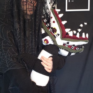 Puede incluir: Una persona con un velo de encaje negro y un su&eacute;ter negro sostiene una criatura de crochet multicolor, a rayas y con patrones. La criatura tiene una boca grande con dientes y un cuerpo largo y delgado. El letrero detr&aacute;s de la persona dice "Beetlejuice" dos veces.