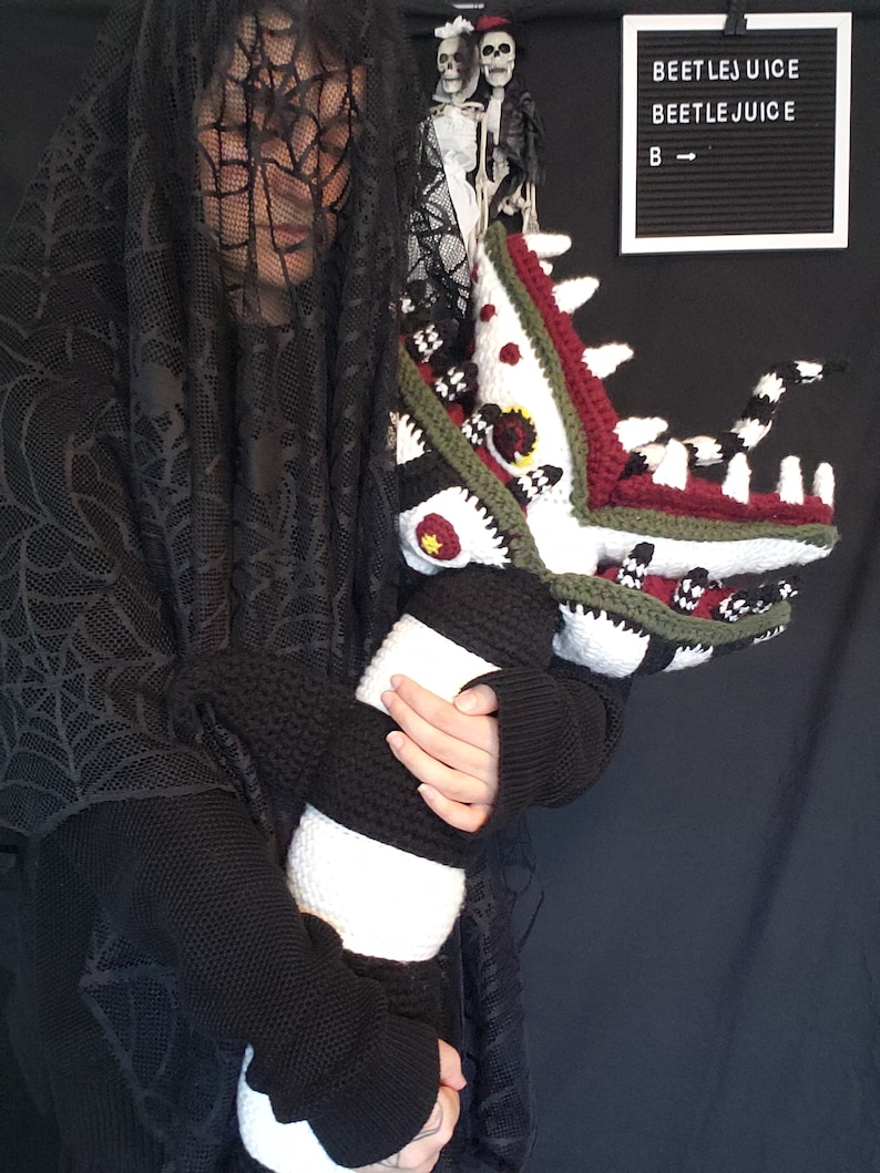 Puede incluir: Una persona con un velo de encaje negro y un su&eacute;ter negro sostiene una criatura de crochet multicolor, a rayas y con patrones. La criatura tiene una boca grande con dientes y un cuerpo largo y delgado. El letrero detr&aacute;s de la persona dice "Beetlejuice" dos veces.