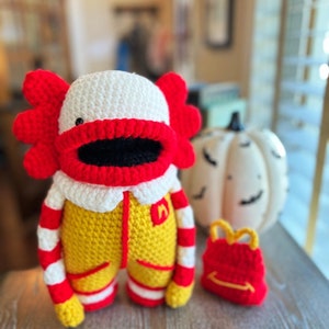 May include: A crocheted clown doll wearing a yellow and red jumpsuit with a white collar and a red and white striped sleeve. The doll is holding a red backpack with a yellow 'M' logo. The doll is sitting on a wooden surface next to a white pumpkin.