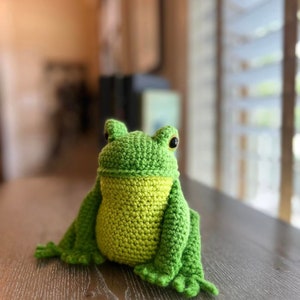 May include: A green crocheted frog toy with yellow accents and black eyes. The frog is sitting on a wooden surface.