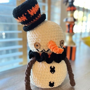 May include: A crocheted snowman wearing a black and orange hat with a brown crocheted scarf and two black spider accents on its body.