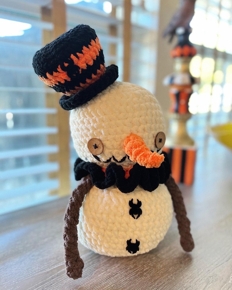 May include: A crocheted snowman wearing a black and orange hat with a brown crocheted scarf and two black spider accents on its body.