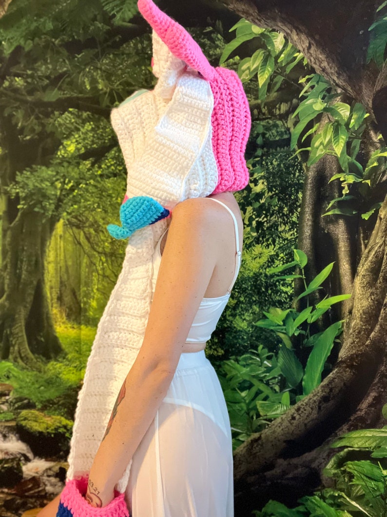 May include: A handmade crochet unicorn hat with a long white scarf, pink horn and ear details, and a blue and pink accent. The hat is part of a costume or accessory set.