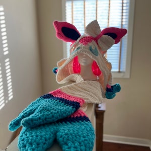 May include: A crochet hat and scarf set with a fantasy theme. The hat has large pink and blue ears, a white hood, and blue details. The scarf and tail are in shades of blue, pink, and white, creating a playful accessory. The set is handmade.