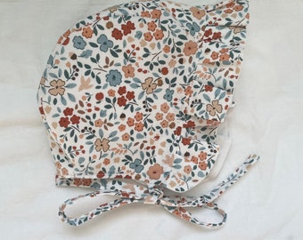 May include: A white bonnet with a ruffled edge and a floral print featuring small blue, brown, and orange flowers. The bonnet has a tie that can be tied under the chin.