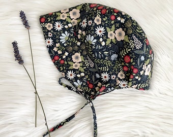 May include: A black fabric bonnet with a floral print featuring white, red, and green flowers. The bonnet has a tie closure.