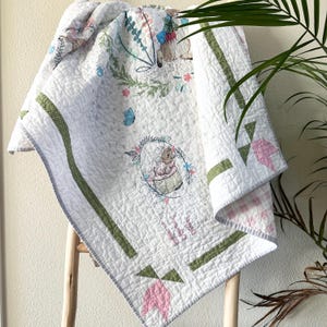 Könnte beinhalten: Weiß gesteppte Babydecke mit Blumen- und Hasenmuster. Die Decke hat grüne und rosa Akzente, mit einem gestickten Bild eines Charakters in einem Blumenkranz. Die Decke ist über eine Holzleiter drapiert.