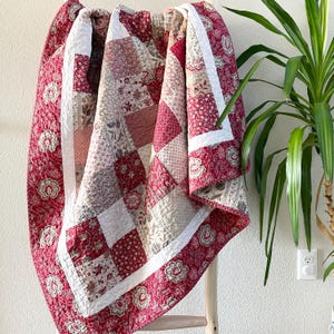 Peut inclure: Une courtepointe rouge et blanche en patchwork avec un motif floral. La courtepointe est pliée et drapée sur une échelle en bois.