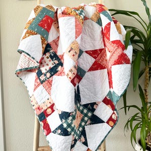 Puede incluir: Un edredón de retazos colorido con una variedad de patrones y colores, incluyendo blanco, rojo, verde azulado y diseños florales. El edredón está drapeado sobre una escalera de madera, mostrando sus intrincadas costuras y diseño. Los bordes del edredón están acabados con un borde verde azulado.