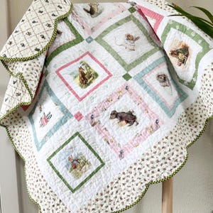 May include: A white quilt with a patchwork design featuring squares with animal illustrations in pink and green frames. The quilt has a scalloped edge with a floral pattern and a green trim.