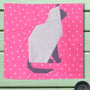 May include: A pink fabric square with a white cross pattern. A gray and black fabric cat shape is layered on top of the pink fabric.