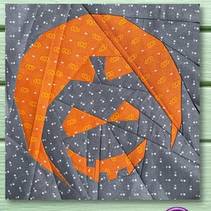 Puede incluir: Un bloque de colcha de retazos de tela gris y naranja con un diseño de calabaza de Halloween. El bloque está formado por pequeños cuadrados de tela cosidos juntos para crear un patrón geométrico.