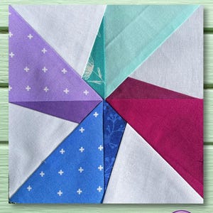 May include: A patchwork quilt block with a pinwheel design. The block is made of eight triangles in various colors, including purple, blue, white, teal, and burgundy. The block is on a light green wooden surface.