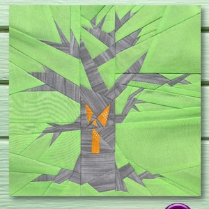 May include: A green fabric patchwork quilt block featuring a grey tree in the centre. The tree has a bright orange window in the trunk. The block is on a light green wooden surface.