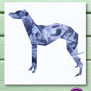 Op de afbeelding: Een witte kaart met een grijs origami-windhond silhouet erop. De kaart ligt op een groene houten ondergrond met roze en grijze knopen en een spoel roze garen.