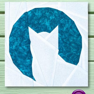 May include: A white fabric square with a teal blue fabric cat shape in the center. The fabric is pieced together in a geometric pattern. The square is on a green wooden surface.