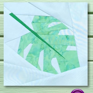 Puede incluir: Un bloque de tela de papel picado verde y blanco con un diseño de hoja de monstera grande. El bloque está sobre una superficie de madera verde claro.