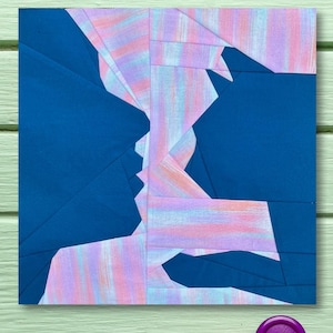 Puede incluir: Una obra de arte abstracta cuadrada que presenta dos perfiles en tonos azules, rosas y blancos. La obra está compuesta por formas y patrones geométricos. Botones rosas y morados, junto con un carrete de hilo rosa, se colocan sobre una superficie de madera verde claro.