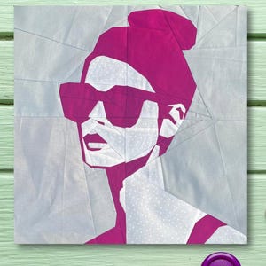 May include: A geometric artwork depicting a woman with a bun and sunglasses in pink and white. The art is set against a light grey backdrop, with a light green wooden surface. Pink and purple buttons and pink thread are scattered around the artwork.