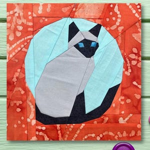 May include: A patchwork quilt block depicting a Siamese cat within a circle, against a light blue background and an orange border. The cat is constructed from different fabric pieces in shades of grey, black, and white.