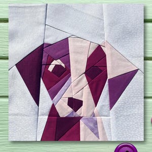 May include: A patchwork quilt block featuring a geometric design of a dog's face. The dog's face is made up of various shades of purple and pink fabric. The background is a light blue fabric.