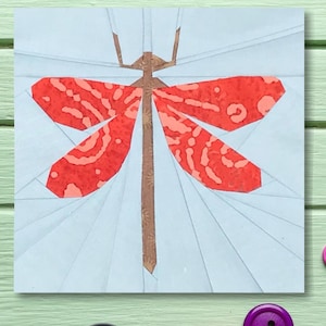 Könnte beinhalten: Ein quadratisches, hellblaues Stoffkunstwerk mit einer stilisierten Libelle mit roten und pfirsichfarbenen Flügeln. Die Libelle hat einen braunen Körper und Kopf. Das Kunstwerk befindet sich auf einer hellgrünen Holzoberfläche mit Knöpfen und rosa Faden.
