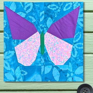 May include: A square fabric block featuring a colorful butterfly design. The butterfly has purple wings, pink and white speckled lower wings, and a green body. The background is a blue batik pattern.