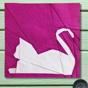 May include: A square, quilted fabric block featuring a white origami-style cat on a magenta background with a small dot pattern. The cat is lying down with its tail curved upwards. The block has a stitched border.