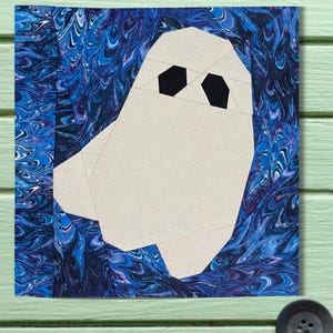 May include: A square fabric art piece depicting a geometric ghost. The ghost is constructed from light beige fabric with black hexagonal eyes, set against a backdrop of swirling blue and white patterned fabric. The artwork resembles a quilt block.