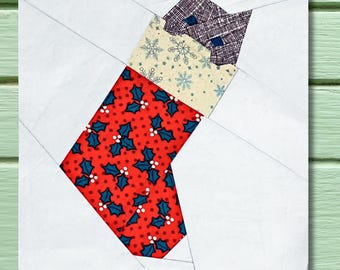 Op de afbeelding: Een stoffen kerstsokken quiltblok. De sok heeft een rood hulstbessenpatroon, een crèmekleurig gedeelte met sneeuwvlokken en een paarse patroon bovenkant. De achtergrond is wit met diagonale lijnen.