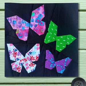 May include: A square, black fabric piece featuring four colorful butterfly designs. Each butterfly is made from patterned fabric in shades of pink, green, purple, and white, with a variety of floral and polka dot patterns. The butterflies are arranged in a symmetrical layout.