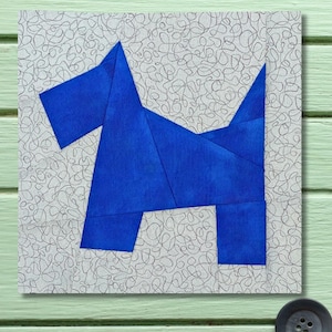 May include: A square fabric block featuring a geometric design of a blue dog against a light gray background with a scribbled pattern. The dog is constructed from angular blue fabric pieces. The block is set against a light green wooden surface.