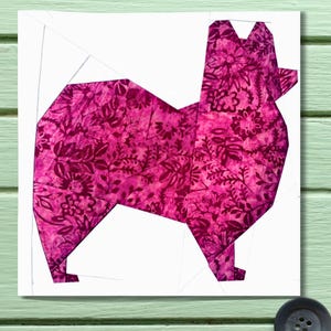 Pode incluir: Um design geométrico de um cão em tons de rosa e magenta, com um padrão floral. O cão está sobre um fundo branco, criando um contraste marcante. A obra de arte é provavelmente um bloco de colcha ou um projeto de artesanato semelhante.