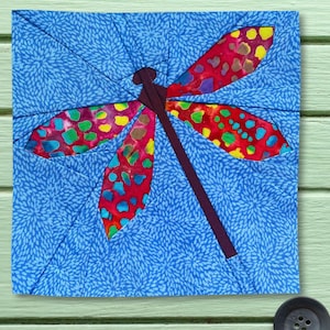 Op de afbeelding: Een vierkant stoffen quiltblok met een libelleontwerp. De libelle heeft een donkerpaars lichaam en vier vleugels van rode stof met een veelkleurig vlekkenpatroon. De achtergrond is blauwe stof met een herhalende bloemenprint.
